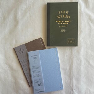 May include: Three notebooks on a white surface. The top notebook is olive green with gold text that reads "LIFE KLEID NOBLE NOTE SECTION 100 SHEETS." Two smaller notebooks are light blue and brown. The notebooks are labeled "KLEID NOTES."