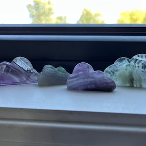 May include: Four small, carved fluorite cloud sculptures in shades of purple, green, and white. The clouds are arranged in a row on a white surface.