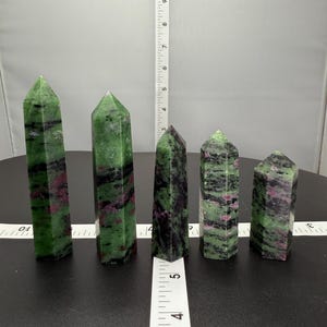 May include: Five green, black, and pink gemstone points, also known as zoisite, arranged in a row on a black surface with a white ruler for scale. The points range in size from approximately 5 cm to 10 cm in length.