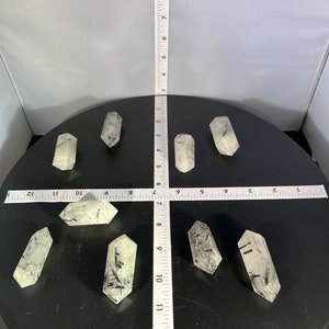 May include: Eight clear quartz crystal points with black tourmaline inclusions. The points are arranged in a circle on a black surface.