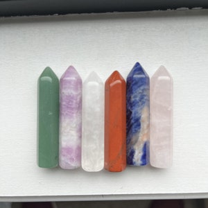 May include: Seven crystal wands in various colors, including green, purple, white, red, blue, and pink. The wands are arranged in a row on a white surface.