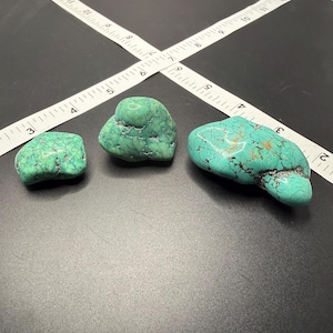 May include: Three turquoise stones with natural black matrix veining, arranged in a row. The stones are positioned next to a white measuring tape showing imperial units, with the largest stone measuring approximately 2.5 inches in length.