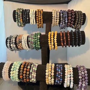 May include: A display of 20 colorful gemstone bracelets on a black stand. The bracelets are made of various stones, including amethyst, agate, aventurine, and tiger's eye.