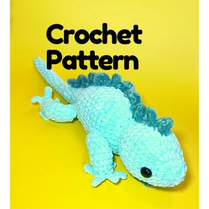 May include: Crochet pattern for a light blue iguana with a dark green spiky back. The iguana has black eyes and is lying on a yellow surface.