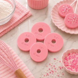 May include: Four circular pink silicone spacers with center holes, each embossed with specific measurements: 1 inch (25 mm), 3/4 inch (19 mm), 5/8 inch (16 mm), and 1/2 inch (13 mm). These flat, round tools are designed for consistent dough rolling.