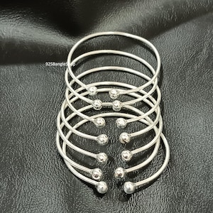 Op de afbeelding: Een stapel zilverkleurige armbanden. Elke armband heeft een gladde, gebogen band die eindigt in een kleine, ronde bal. De armbanden liggen op een zwarte ondergrond. De tekst "925BanglesShop" is zichtbaar.