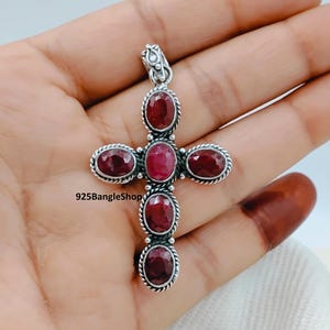 May include: A silver cross pendant featuring oval, deep red gemstones. The gemstones are set in a silver frame with a twisted rope design. The pendant has a loop at the top for a chain. The text "925BangleShop" is visible.