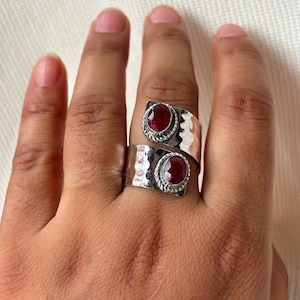 May include: Two silver rings with hammered textures and oval, deep red gemstones. The rings are worn on a hand, showcasing the intricate metalwork and the vibrant color of the stones. The design is a wrap-around style.