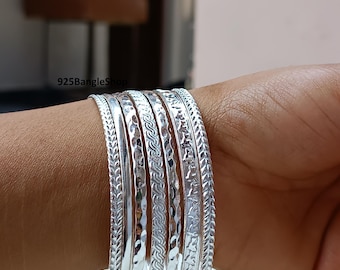Pode incluir: Uma coleção de pulseiras de prata com vários designs em relevo, incluindo padrões florais, geométricos e texturizados. As pulseiras estão empilhadas em um pulso, mostrando seus diferentes estilos e brilho. O nome da loja "925BangleShop" é visível.