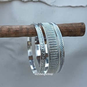 Op de afbeelding: Drie zilveren armbanden met verschillende gegraveerde ontwerpen. De armbanden zijn gestapeld op een houten stok. De tekst "925BangleShop" is zichtbaar onderaan de afbeelding.
