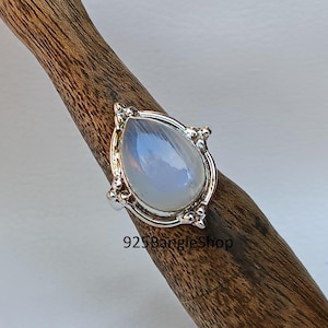 May include: A silver ring featuring a teardrop-shaped moonstone set in an ornate silver setting. The ring is displayed on a wooden surface.