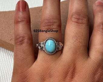 Può includere: Un anello d'argento con una pietra ovale azzurra. L'anello presenta un motivo a corda intrecciata attorno alla pietra e volute decorative sui lati. Il testo "925BangleShop" è visibile.