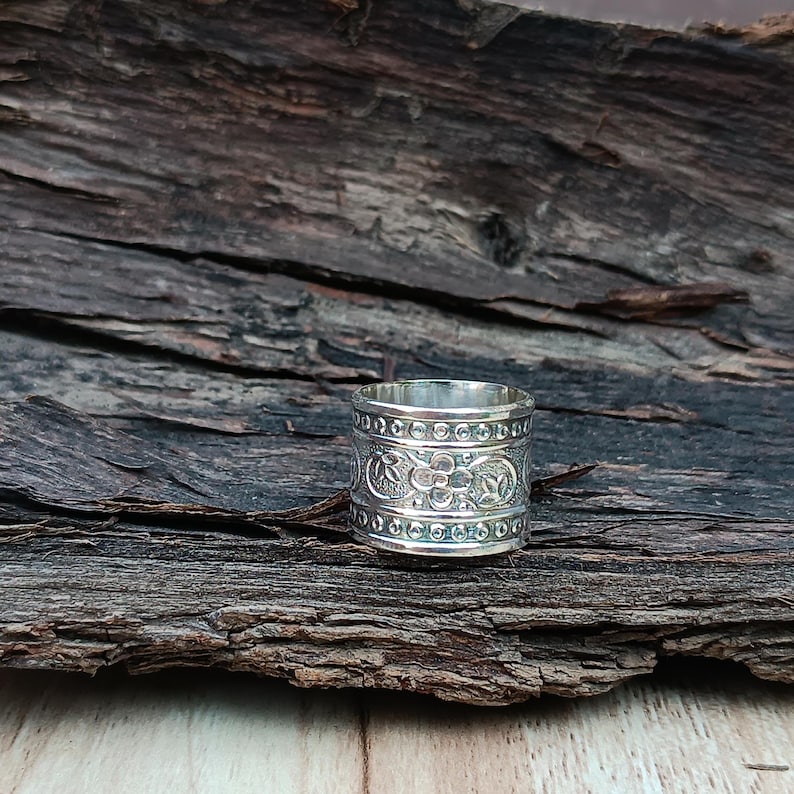 K&ouml;nnte beinhalten: Ein silberner Ring mit einem breiten Band, das mit einem komplizierten Blumenmuster verziert ist. Der Ring liegt auf einem rustikalen Holzgrund.