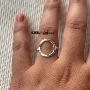 Puede incluir: Un anillo de plata con un diseño de círculo abierto. El anillo se lleva en un dedo. El texto "925BangleShop" es visible en la imagen.