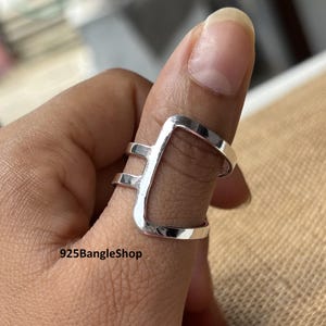 May include: A silver ring with a geometric design is worn on a finger. The ring has a rectangular shape with two parallel bars. The text "925BangleShop" is visible at the bottom of the image.