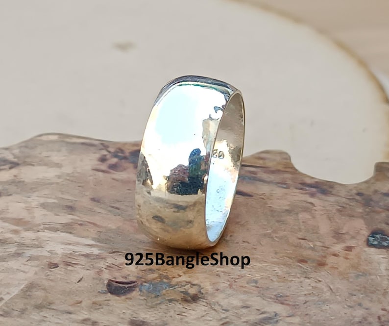 May include: A polished silver ring with a rounded band. The ring reflects the surrounding environment, creating a mirrored effect. The text "925BangleShop" is visible at the bottom of the image. The ring is likely made of sterling silver.