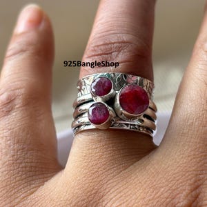 Può includere: Un anello d'argento con tre grandi pietre preziose rotonde di colore rosso intenso. L'anello ha una fascia strutturata e viene indossato su un dito. Il testo "925BangleShop" è visibile sull'anello.