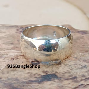 May include: A wide, polished silver ring with a smooth, rounded design. The ring reflects the surrounding environment, creating a mirrored effect. The ring is displayed on a textured, brown surface. The text "925BangleShop" is visible at the bottom.