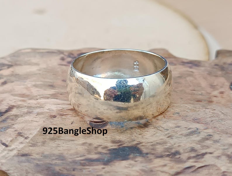 May include: A wide, polished silver ring with a smooth, rounded design. The ring reflects the surrounding environment, creating a mirrored effect. The ring is displayed on a textured, brown surface. The text "925BangleShop" is visible at the bottom.