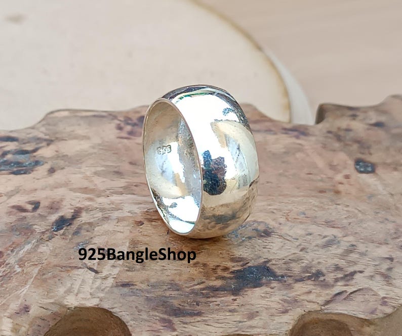 May include: A wide, polished silver ring. The ring has a smooth, rounded design and a bright, reflective surface. The text "925BangleShop" is visible at the bottom of the image. The ring is displayed on a wooden surface.