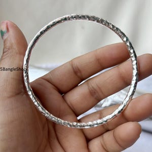 Puede incluir: Un brazalete de plata con textura martillada. El brazalete redondo se sostiene en una mano, mostrando su superficie reflectante. El brazalete es una pieza de joyería, probablemente usada como accesorio.