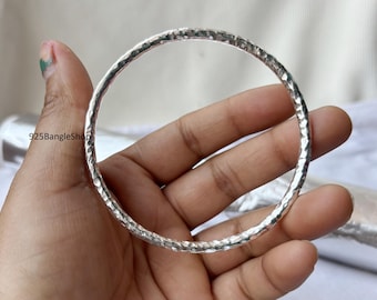 May include: A silver bangle with a hammered texture. The round bracelet is held in a hand, showcasing its reflective surface. The bangle is a piece of jewelry, likely worn as an accessory.