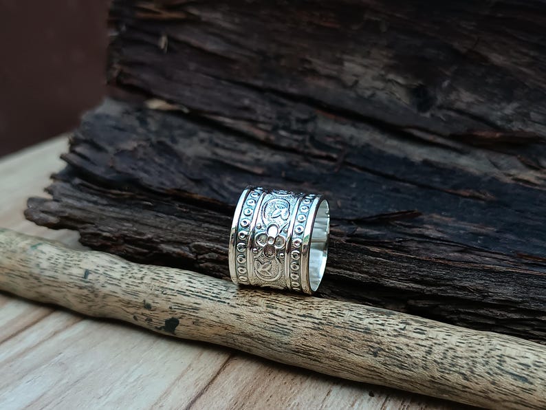 K&ouml;nnte beinhalten: Ein silberner Ring mit einem dekorativen Blumenmuster. Der Ring ist breit und hat eine glatte, polierte Oberfl&auml;che.