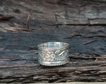 May include: A silver ring with an engraved floral and leaf pattern. The ring has a band design with a detailed floral motif in the center, surrounded by beaded and leaf accents. The ring is displayed on a dark wooden surface.