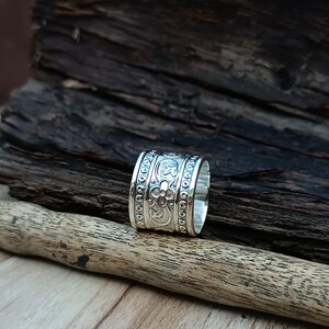 K&ouml;nnte beinhalten: Ein silberner Ring mit einem dekorativen Blumenmuster. Der Ring ist breit und hat eine glatte, polierte Oberfl&auml;che.