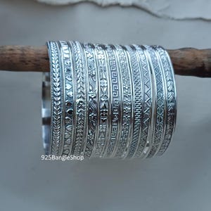 May include: A stack of 12 silver bangles with different engraved designs. The bangles are stacked on a wooden surface. 925BangleShop is printed on the bottom of the image.