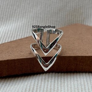 May include: A silver ring featuring a double V-shaped design. The ring is made of shiny metal and is displayed on a brown surface. The text "925BangleShop" is visible in the image.