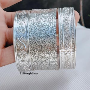 Puede incluir: Tres pulseras de plata con intrincados diseños florales y de volutas. Las pulseras se muestran sobre un fondo neutro, mostrando los patrones y texturas detallados. El texto "925BangleShop" es visible.