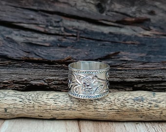 May include: A silver ring with a floral design. The ring is wide and has a textured surface. The ring is sitting on a piece of driftwood.
