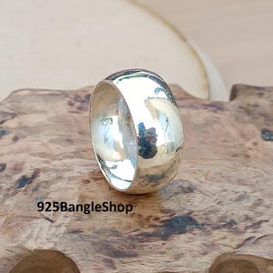 May include: A wide, polished silver ring. The ring has a smooth, rounded design and a bright, reflective surface. The text "925BangleShop" is visible at the bottom of the image. The ring is displayed on a wooden surface.