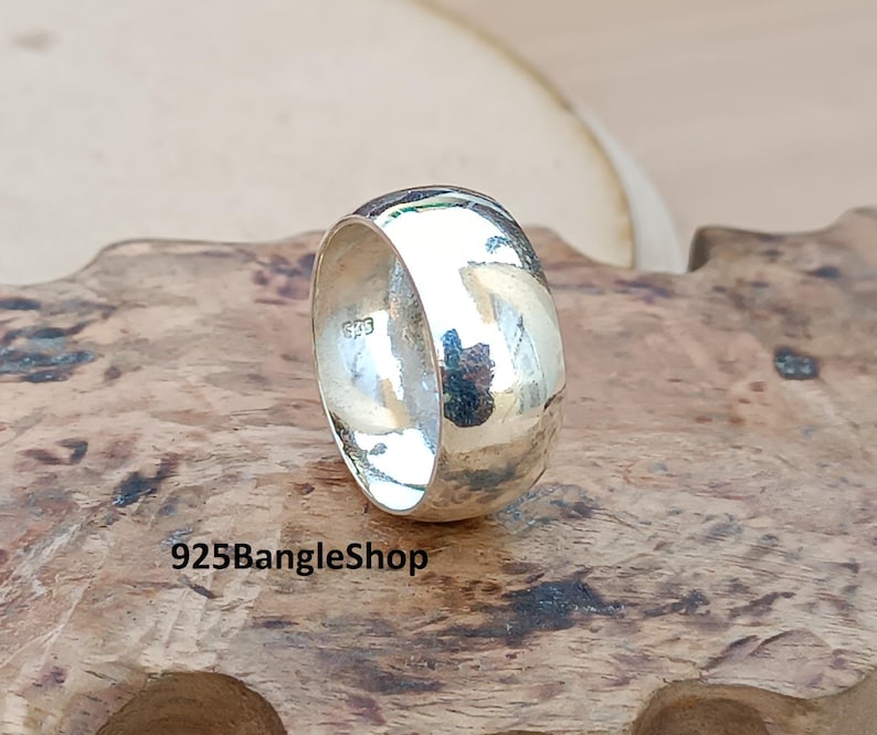 May include: A polished silver ring with a wide band. The ring is smooth and reflective, with the text "925" visible on the inside. The ring is displayed on a wooden surface. The shop name "925BangleShop" is visible.