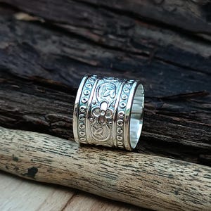 K&ouml;nnte beinhalten: Ein silberner Ring mit einem breiten Band, das mit einem komplizierten floralen Muster verziert ist. Das Band ist mit einem sich wiederholenden Muster aus Blumen und Bl&auml;ttern verziert.