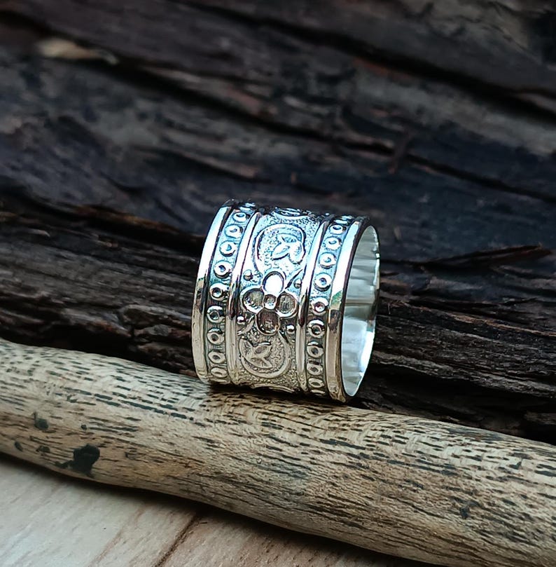 K&ouml;nnte beinhalten: Ein silberner Ring mit einem breiten Band, das mit einem komplizierten floralen Muster verziert ist. Das Band ist mit einem sich wiederholenden Muster aus Blumen und Bl&auml;ttern verziert.