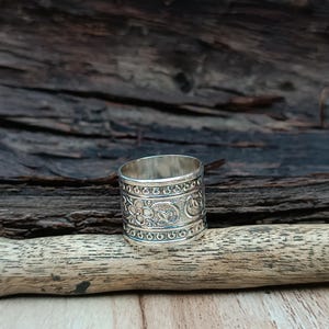 K&ouml;nnte beinhalten: Ein silberner Ring mit einem komplizierten floralen und geometrischen Design. Der Ring ist breit und hat eine polierte Oberfl&auml;che.