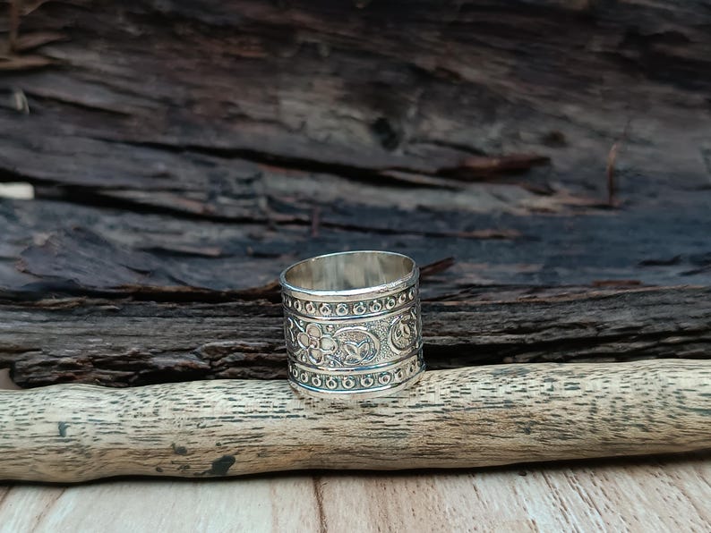 K&ouml;nnte beinhalten: Ein silberner Ring mit einem komplizierten floralen und geometrischen Design. Der Ring ist breit und hat eine polierte Oberfl&auml;che.