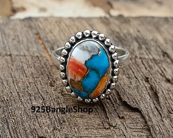 May include: A silver ring with an oval-shaped stone featuring a mix of colors: white, orange, and turquoise. The stone is surrounded by a beaded silver border. The text "925BangleShop" is visible.
