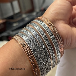 Op de afbeelding: Een verzameling zilver- en koperkleurige armbanden met ingewikkelde bloemen- en bladerpatronen. De armbanden zijn op een pols gestapeld, waarbij de gedetailleerde ontwerpen en texturen worden getoond. De tekst "925BangleShop" is zichtbaar.