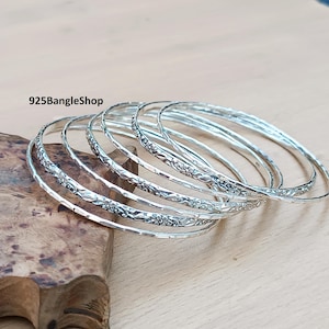 Può includere: Un gruppo di braccialetti d'argento con motivi floreali. I braccialetti sono esposti su una superficie di legno. Il testo "925BangleShop" è visibile nell'immagine.