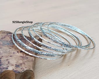 May include: A collection of silver bangles with floral detailing. The bangles are presented on a wooden surface. The text "925BangleShop" is visible in the image.