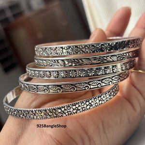 Op de afbeelding: Een verzameling zilveren armbanden met ingewikkelde bloemen- en bladerpatronen. De armbanden worden op een hand getoond, waarbij de gedetailleerde ontwerpen worden getoond. De tekst "925BangleShop" is zichtbaar op een van de armbanden.