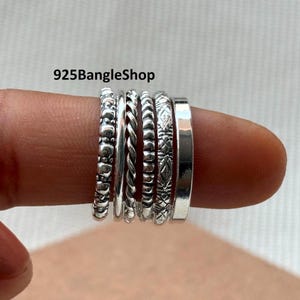 May include: A collection of silver rings with various designs, including beaded, twisted, and patterned bands. The rings are stacked on a finger, showcasing their different textures and styles. The shop name "925BangleShop" is visible.
