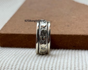 Può includere: Un anello color argento con una fascia rotante con ritagli a forma di cuore. L'anello ha un design strutturato e il testo "925BangleShop" è visibile. L'anello è esposto su una superficie bianca.