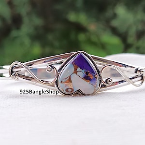 May include: A silver bangle bracelet with a heart-shaped stone. The stone is a mix of purple, white, and gold. The bracelet has decorative swirls on each side of the heart. The text "925BangleShop" is visible.