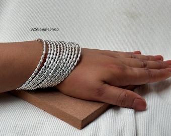 May include: A collection of silver-coloured, twisted-rope design bangles. The bangles are stacked on a person's wrist. The image is set against a neutral background. The shop name "925BangleShop" is visible in the image.