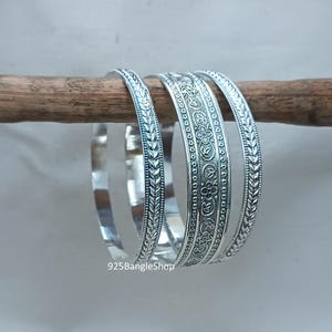 Op de afbeelding: Vier zilveren armbanden met gedetailleerde patronen worden getoond op een houten deuvel. Twee armbanden hebben een bladerenpatroon en twee hebben een bloemenmotief. De armbanden zijn gemaakt van glanzend metaal en staan tegen een neutrale achtergrond.