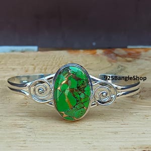 May include: A silver bangle bracelet featuring a green and copper oval turquoise stone set in the centre. The bracelet has a swirl design on each side of the stone.
